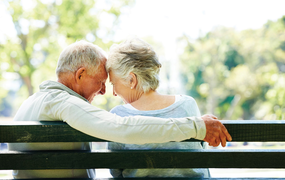 Two residents enjoying romantic moment on park bench in garden at assisted living community.